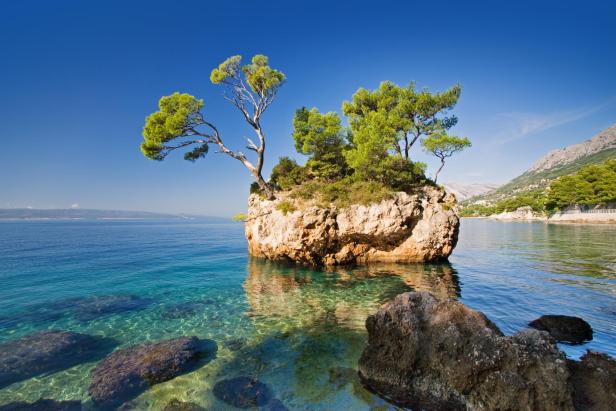 Eine kleine, bewaldete Insel im klaren, türkisfarbenen Wasser vor einer Küstenlandschaft.