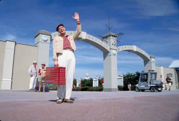 Jim Carrey als Truman aus dem Film „Truman Show“. Sein Charakter steht mit Koffer winkend auf rotem Pflaster und winkt, weil er verreisen will.