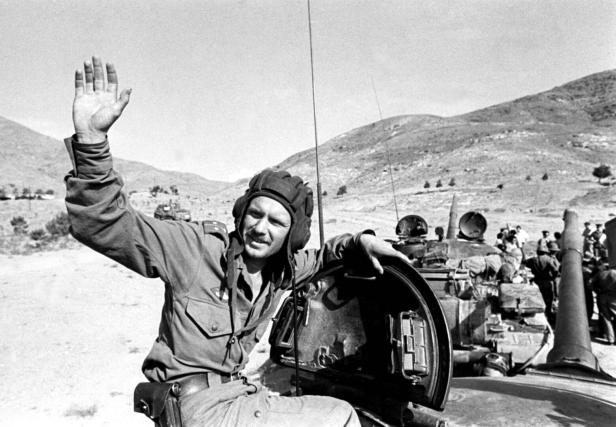Ein Panzerkommandant winkt von seinem Panzer in einer hügeligen Landschaft.