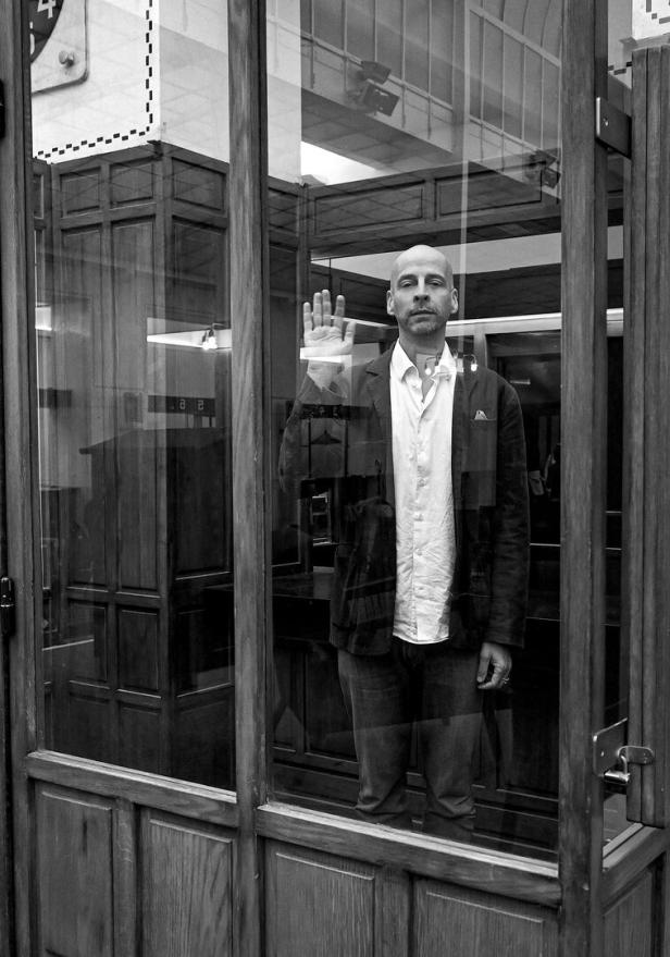 Paul Divjak steht hinter einer Glasscheibe und hebt die Hand zu einer Winkbewegung.