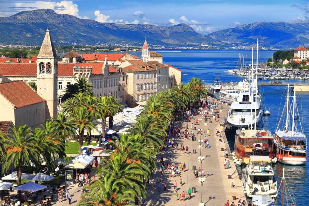 Blick auf die Uferpromenade von Trogir, Kroatien, mit Palmen, Menschen und Booten.
