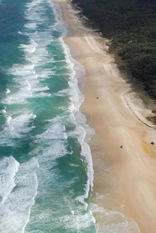Ein Sandstrand mit türkisfarbenem Wasser und einigen Fahrzeugen aus der Vogelperspektive.