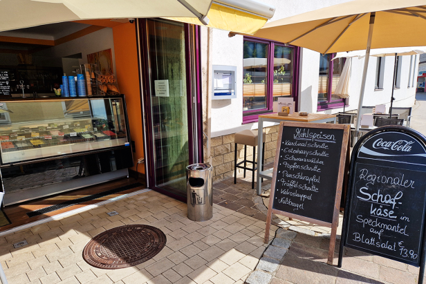 Einladende Außensicht eines Cafés mit Tischen, Sonnenschirmen und einer Tafel mit Mehlspeisen.