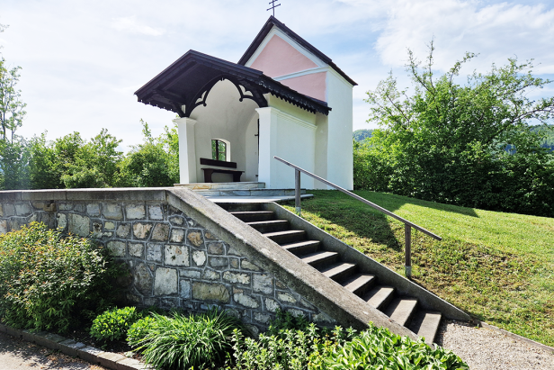 Eine kleine Kapelle mit Treppe steht auf einer grünen Anhöhe.