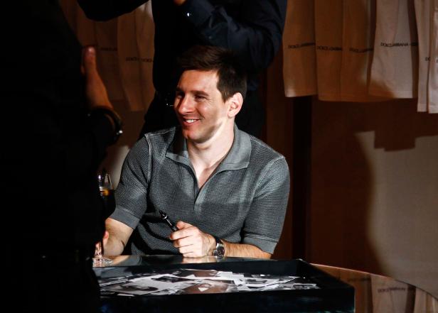 Lionel Messi sitzt lächelnd an einem Tisch und signiert Fotos.