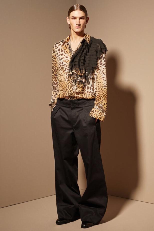Ein Mann in einer Leopardenmusterbluse mit Rüschen und einer weiten schwarzen Hose posiert.