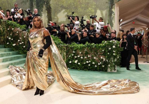 Serena Williams in einem goldenen Kleid auf dem grünen Teppich, umgeben von Fotografen.