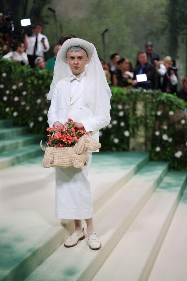 Ein Model in einem weißen Anzug, Hut mit Schleier und einer Dackel-Handtasche voller Blumen.