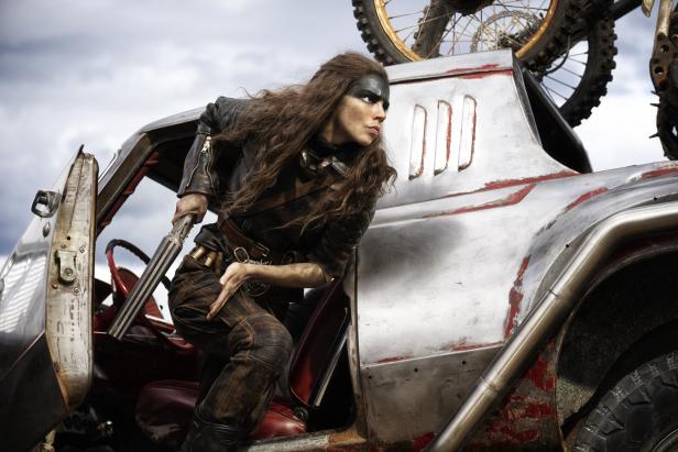 Furiosa springt mit einer abgesägten Schrotflinte aus einem gepanzerten Wagen. Sie trägt eine Lederjacke und hat ihr Gesicht schwarz angemalt.