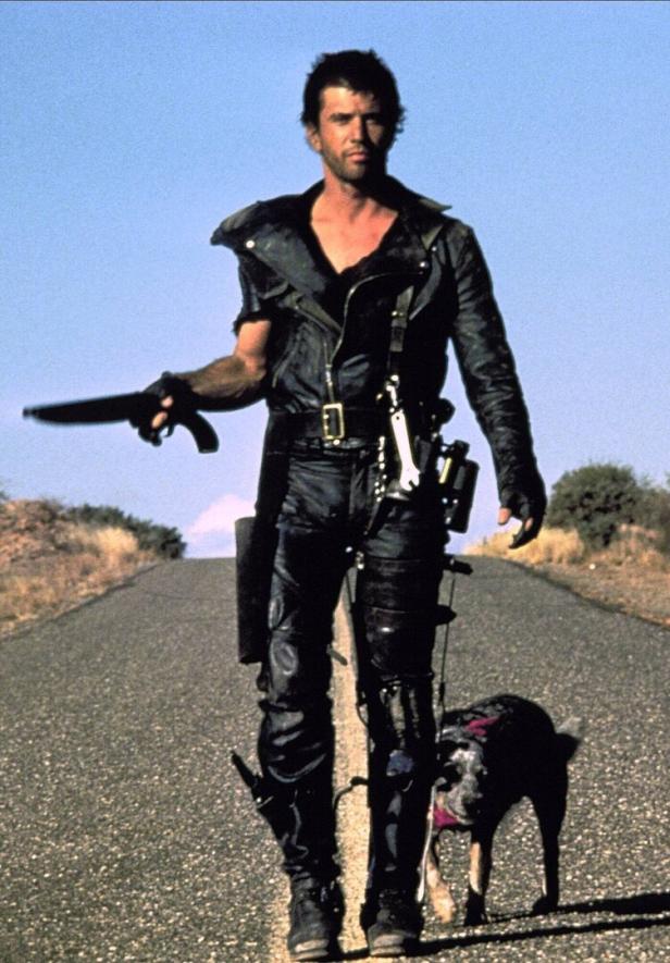 Mel Gibson im Film „Mad Max 2“: Er trägt schwarze Motorradkleidung aus Leder, hält eine Pistole in der Hand und ein Hund trottet hinter ihm in der Wüste.