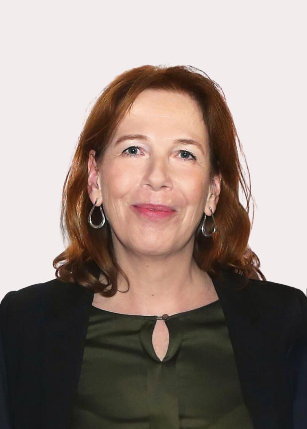 Porträt einer Frau mit roten Haaren, die eine grüne Bluse und einen schwarzen Blazer trägt.
