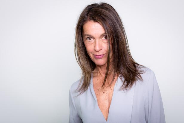 Porträt einer Frau mit braunen Haaren und einem hellblauen Blazer vor einem weißen Hintergrund.