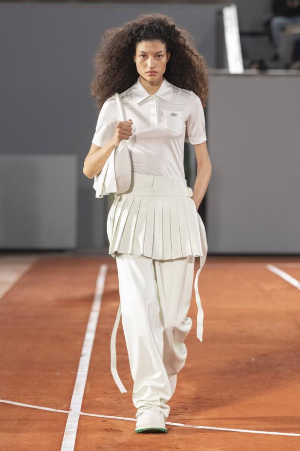 Eine Frau mit lockigem Haar präsentiert ein weißes Outfit von Lacoste auf einem Laufsteg.
