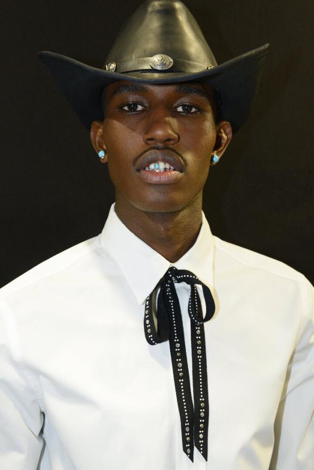Ein Mann mit Cowboyhut, türkisfarbenen Ohrringen und Grillz vor schwarzem Hintergrund.