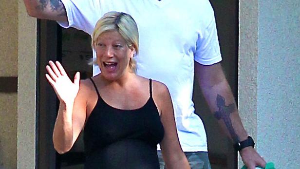 Eine schwangere Frau mit blonden Haaren winkt in die Kamera.