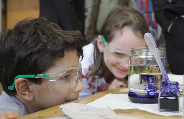 Zwei Kinder mit Schutzbrillen beobachten ein Experiment mit Flüssigkeiten in Gläsern.