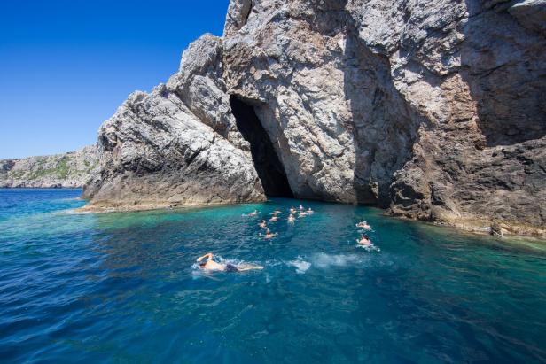 Eine Gruppe von Menschen schwimmt im türkisfarbenen Wasser vor einer felsigen Küste mit einer Höhle.