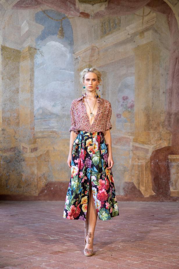 Eine Frau in einem floralen Outfit steht vor einer Wandmalerei.
