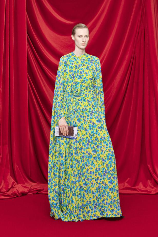 Eine Frau in einem gelb-blauem Blumenkleid hält einen Reisepass und eine Flugticket vor einem roten Vorhang.