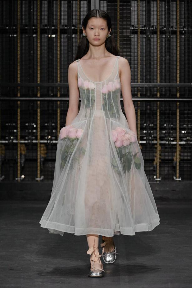 Ein Model präsentiert ein transparentes Kleid mit aufgenähten Rosen auf einem Laufsteg.