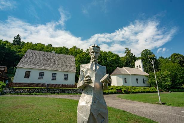 Eine Statue von Nikola Tesla vor einem traditionellen kroatischen Haus und einer Kirche.
