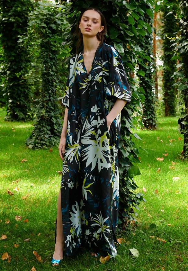 Eine Frau posiert in einem floralen Maxikleid vor einem Baum im Wald.