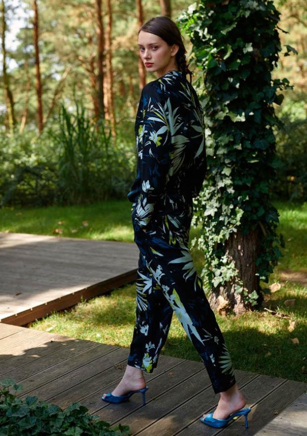 Eine Frau in einem floralen Hosenanzug und blauen Schuhen geht auf einem Holzweg.