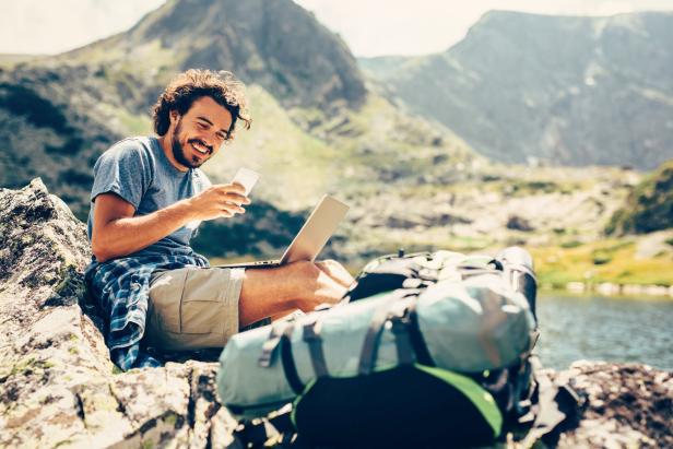 Mann mit Handy, Laptop und Campingrucksack in den Bergen, auf einem Fels sitzend und lachend, im Hintergrund ein Bergsee