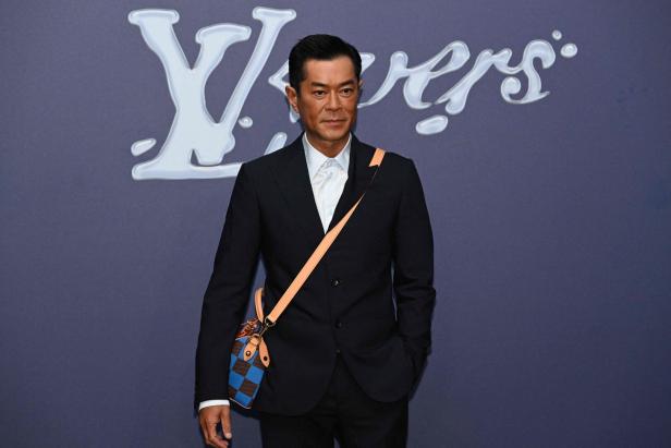 Ein Mann mit Anzug und Umhängetasche posiert vor einem Louis Vuitton-Logo.