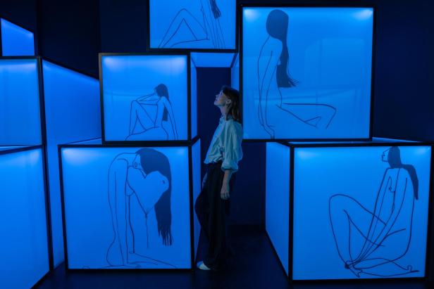 Eine Frau steht in einer Installation aus blau beleuchteten Würfeln mit Frauenskizzen.