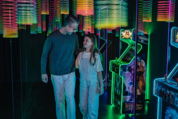 Ein Vater und seine Tochter stehen in einer Spielhalle mit leuchtenden Arcade-Automaten.