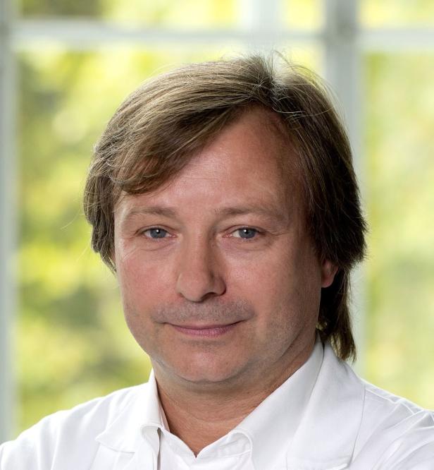 Porträt eines Mannes mit hellbraunen Haaren und einem weißen Hemd vor einem Fenster.