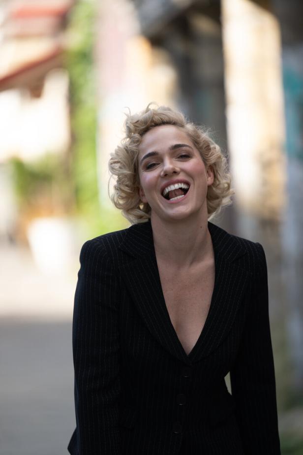 Eine lachende Frau mit blonden Locken trägt einen dunklen Blazer.