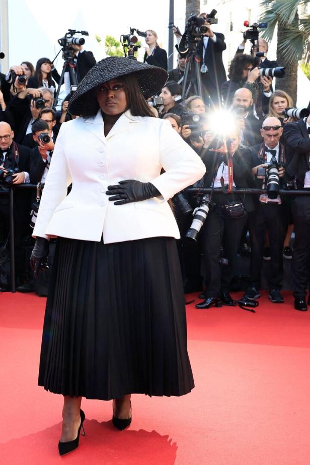 Eine Frau in einem weißen Blazer, schwarzem Rock und Hut posiert auf einem roten Teppich vor Fotografen.