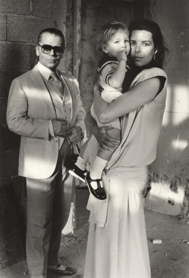 Karl Lagerfeld in den Achtzigern mit Sonnenbrille und Dreiteiler-Anzug. Die junge Caroline von Monaco im Sommerkleid hält ihren Sohn, der eine Matrosenkleidung trägt.
