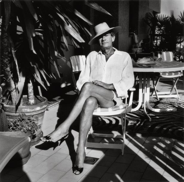 Helmut Newton sitzt auf einer Terrasse. Er hat Pumps an und die Beine überschlagen. Auf seinem Kopf sitzt ein Damenhut.