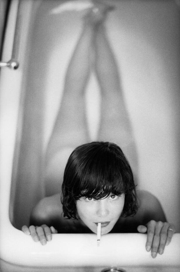 Eine nackte Frau liegt bauchlings in einer Badewanne. Sie hat einen Pagenkopf und eine Zigarette im Mund.