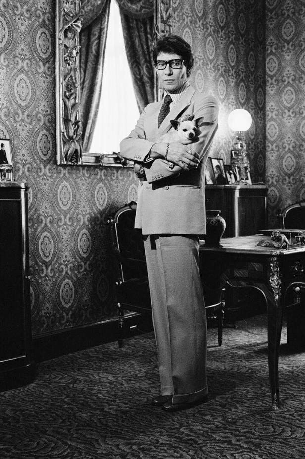 Yves Saint Laurent steht mit Anzug in einem Zimmer mit üppigen Wandtextilien. Er hat einen Hund im Arm.