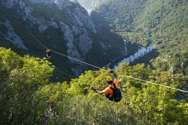 Zwei Personen sausen an einer Zipline über eine bewaldete Schlucht.