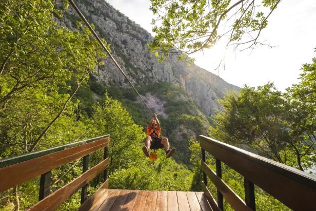 Ein Mann saust an einer Zipline durch eine grüne Berglandschaft.