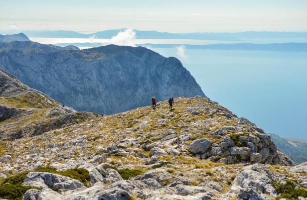Zwei Wanderer auf einem felsigen Berggipfel mit Blick auf das Meer.