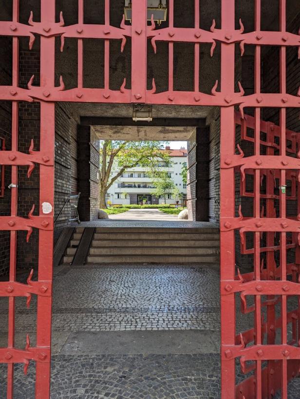 Ein Blick durch ein rotes, offenes Tor auf einen begrünten Innenhof mit Wohnhäusern im Hintergrund.