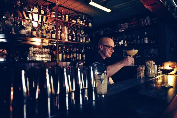 Ein Barkeeper mit Brille serviert einen Cocktail vor einem Regal voller Spirituosen.