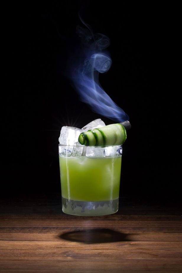 Ein grüner Cocktail mit Eis und einer Gurkengarnitur, von dem Rauch aufsteigt.