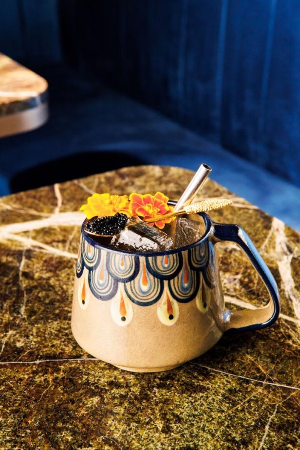 Ein Cocktail in einem verzierten Becher mit Eis, Beeren und Blüten auf einem Tisch.