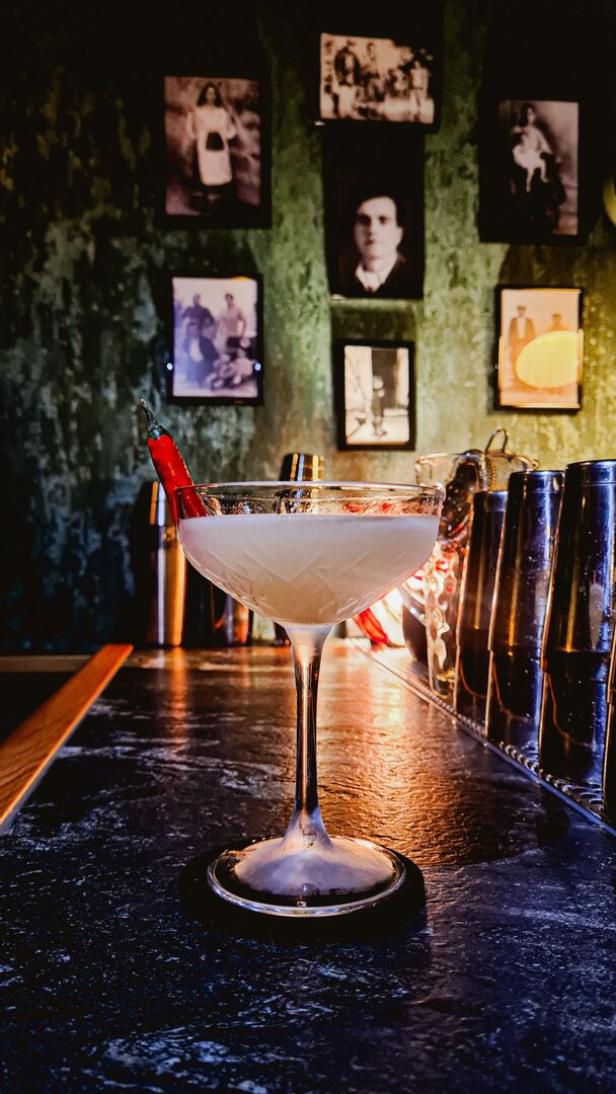 Ein Cocktail in einem eleganten Glas, garniert mit einer roten Chili, steht auf einer Bar.