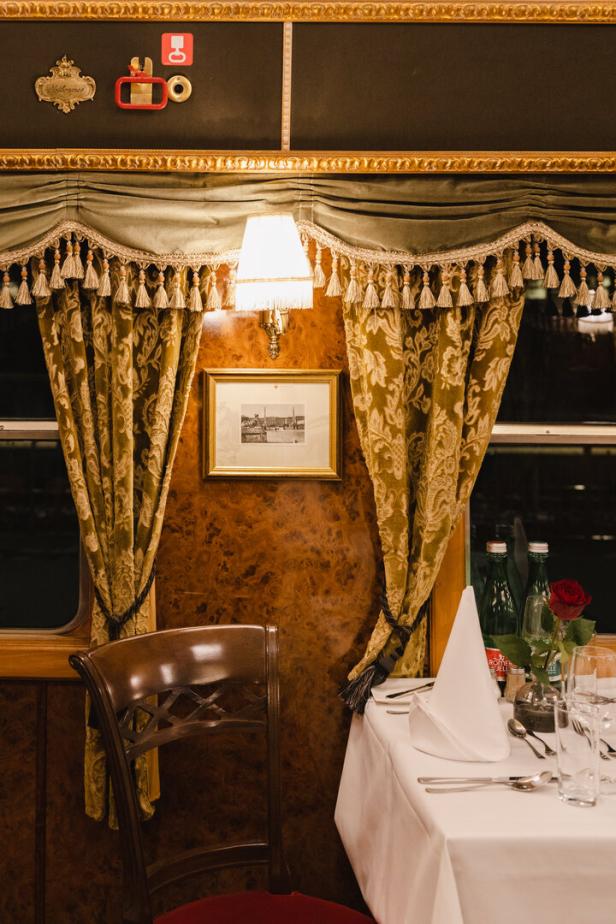 Ein gedeckter Tisch in einem luxuriösen Zugabteil mit Vorhängen und einer Lampe.