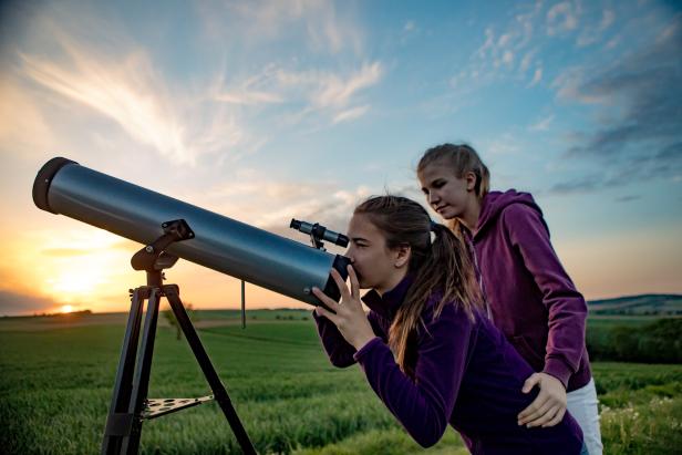 Zwei Mädchen beobachten den Sonnenuntergang durch ein Teleskop auf einem Feld.