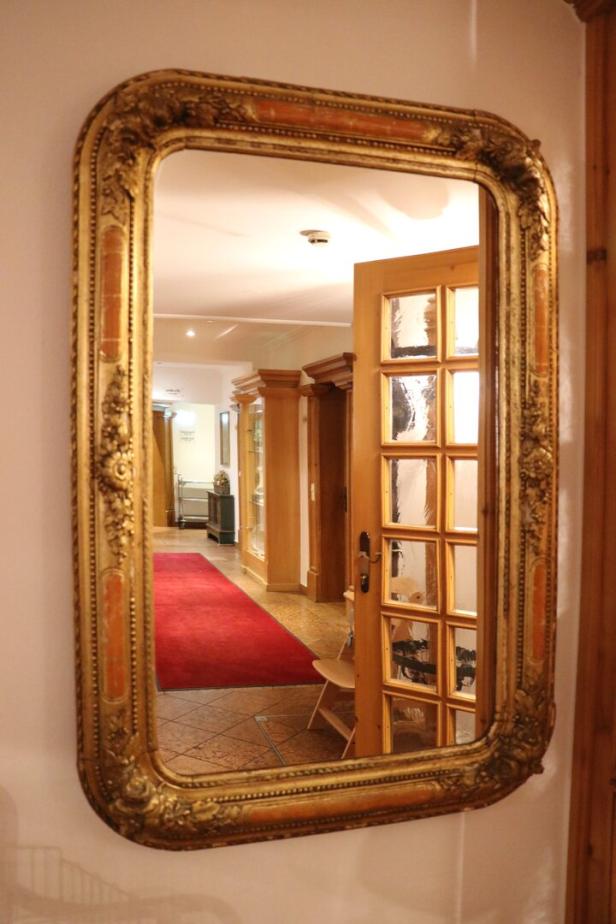 Ein goldener Spiegel reflektiert einen Flur mit einem roten Teppich und einer Holztür.