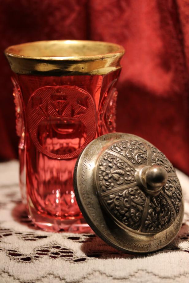 Ein rotes Glasgefäß mit Goldrand und ein verzierter Deckel liegen auf einer gehäkelten Decke.